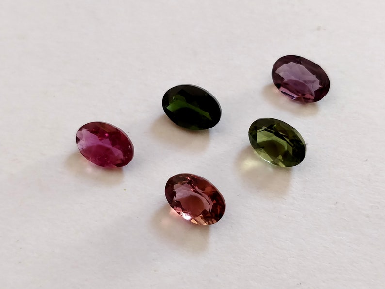 May include: Five oval-shaped gemstones in various colors, including pink, green, and purple. The gemstones are arranged on a white surface.