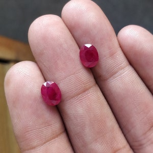 Puede incluir: Dos piedras preciosas de rubí de forma ovalada, cada una de un color rojo intenso, se muestran en la mano de una persona.