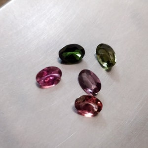 May include: Five oval-shaped gemstones in various shades of green, pink, and purple. The gemstones are arranged in a semi-circle on a silver surface.