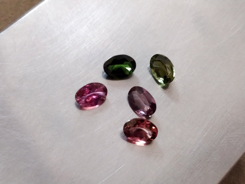 May include: Five oval-shaped gemstones in various shades of green, pink, and purple. The gemstones are arranged in a semi-circle on a silver surface.