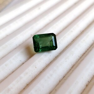 May include: A dark green emerald cut gemstone on a white background.