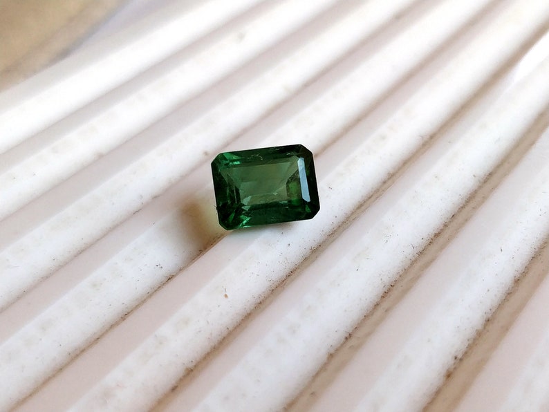 May include: A dark green emerald cut gemstone on a white background.