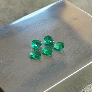 K&ouml;nnte beinhalten: F&uuml;nf lose, birnenf&ouml;rmige Smaragd-Edelsteine mit leuchtend gr&uuml;ner Farbe und facettierten Oberfl&auml;chen. Die Steine sind in einer Gruppe auf einer glatten, geb&uuml;rsteten Metalloberfl&auml;che angeordnet, die das Licht reflektiert und die Klarheit betont.
