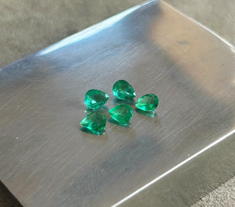 K&ouml;nnte beinhalten: F&uuml;nf lose, birnenf&ouml;rmige Smaragd-Edelsteine mit leuchtend gr&uuml;ner Farbe und facettierten Oberfl&auml;chen. Die Steine sind in einer Gruppe auf einer glatten, geb&uuml;rsteten Metalloberfl&auml;che angeordnet, die das Licht reflektiert und die Klarheit betont.