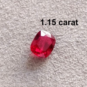 Pode incluir: Uma gema vermelha de 1,15 quilates, provavelmente um rubi, com corte almofada. A gema est&aacute; sobre uma superf&iacute;cie branca texturizada.