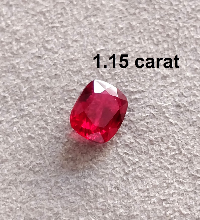 Pode incluir: Uma gema vermelha de 1,15 quilates, provavelmente um rubi, com corte almofada. A gema est&aacute; sobre uma superf&iacute;cie branca texturizada.
