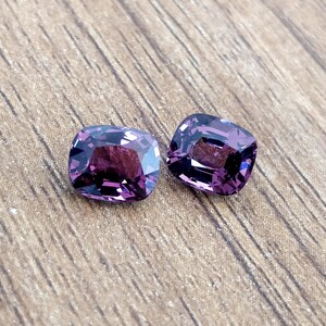 May include: Two purple cushion-cut gemstones on a wooden surface.