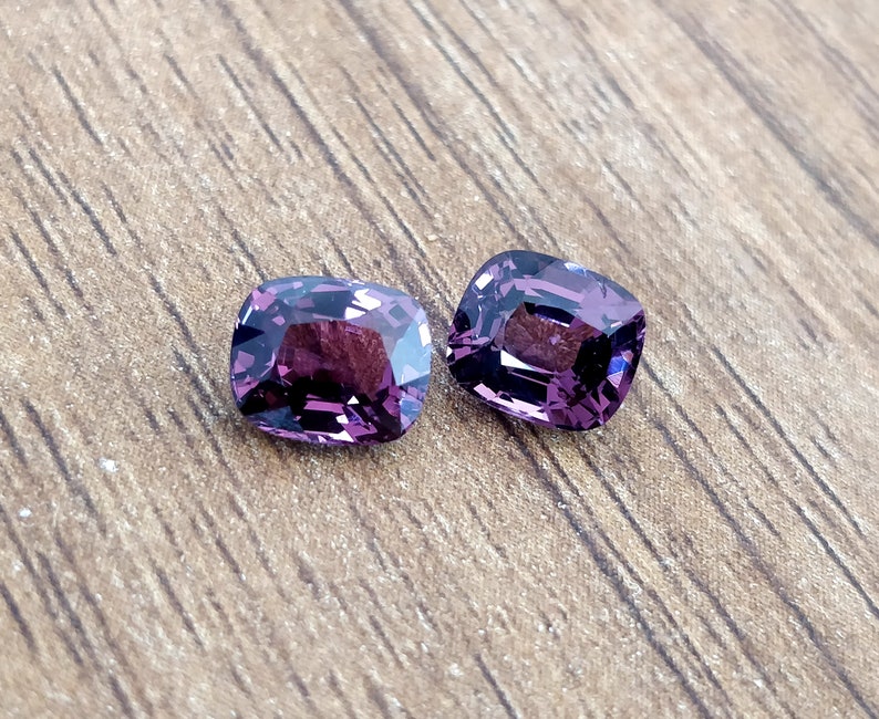 May include: Two purple cushion-cut gemstones on a wooden surface.