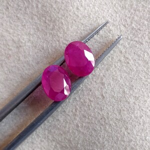 Puede incluir: Dos piedras preciosas ovaladas facetadas de un tono rosa vibrante. Las piedras preciosas est&aacute;n sujetas por unas pinzas sobre un fondo marr&oacute;n claro.
