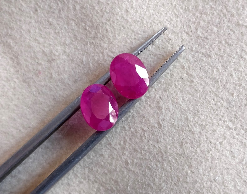Puede incluir: Dos piedras preciosas ovaladas facetadas de un tono rosa vibrante. Las piedras preciosas est&aacute;n sujetas por unas pinzas sobre un fondo marr&oacute;n claro.