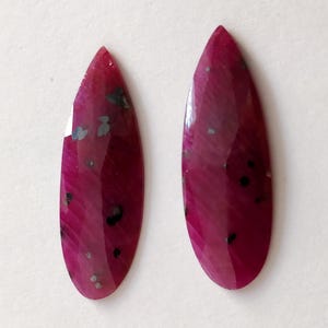 May include: Two pear-shaped ruby gemstones with black and silver inclusions. The stones are a deep pink color and have a smooth, polished finish.