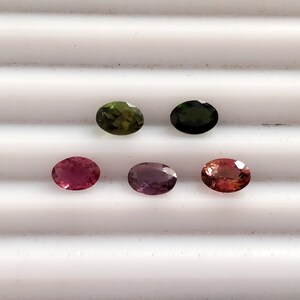 May include: Five oval-shaped gemstones in various shades of green, pink, and purple. The gemstones are arranged in a row on a white surface.