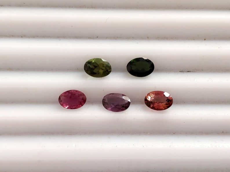 May include: Five oval-shaped gemstones in various shades of green, pink, and purple. The gemstones are arranged in a row on a white surface.