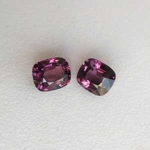 May include: Two cushion-cut gemstones, both a deep purple color, are displayed on a white surface.