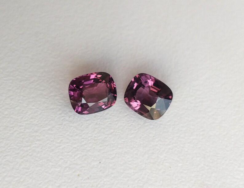 May include: Two cushion-cut gemstones, both a deep purple color, are displayed on a white surface.