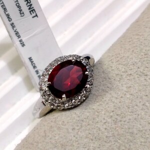 May include: A silver ring with a large oval red gemstone surrounded by smaller white gemstones. The ring is on a gray fabric surface. The tag reads "GARNET (TOPAZ) STERLING SILVER 925 MADE IN"