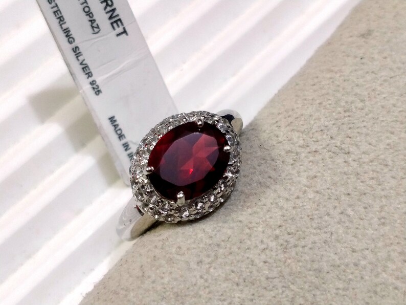 May include: A silver ring with a large oval red gemstone surrounded by smaller white gemstones. The ring is on a gray fabric surface. The tag reads "GARNET (TOPAZ) STERLING SILVER 925 MADE IN"
