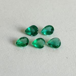 K&ouml;nnte beinhalten: F&uuml;nf lose, birnenf&ouml;rmige Smaragd-Edelsteine mit leuchtend gr&uuml;ner Farbe und facettierten Oberfl&auml;chen. Die Steine sind in einer Gruppe angeordnet und zeigen ihre durchscheinende Qualit&auml;t sowie ihre spitz zulaufenden, tropfenf&ouml;rmigen Silhouetten.