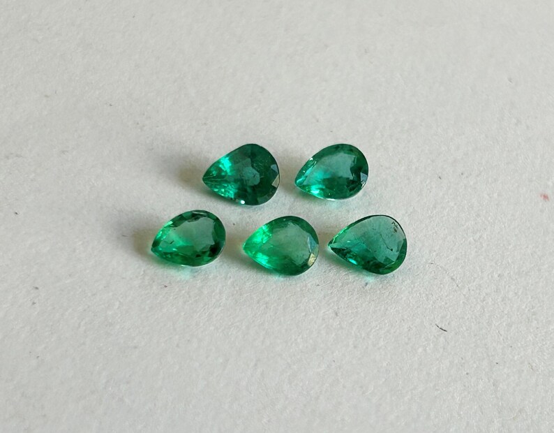 K&ouml;nnte beinhalten: F&uuml;nf lose, birnenf&ouml;rmige Smaragd-Edelsteine mit leuchtend gr&uuml;ner Farbe und facettierten Oberfl&auml;chen. Die Steine sind in einer Gruppe angeordnet und zeigen ihre durchscheinende Qualit&auml;t sowie ihre spitz zulaufenden, tropfenf&ouml;rmigen Silhouetten.