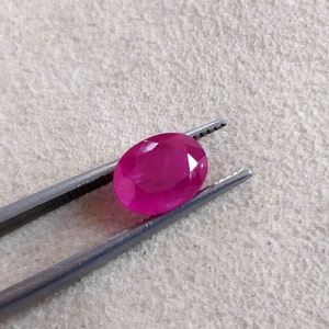 Puede incluir: Una gema ovalada de color rosa vibrante sostenida por unas pinzas. La gema es lisa y pulida, con un ligero brillo.