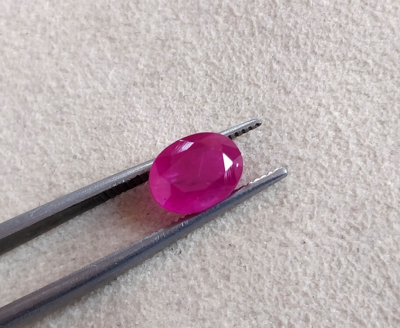 Puede incluir: Una gema ovalada de color rosa vibrante sostenida por unas pinzas. La gema es lisa y pulida, con un ligero brillo.