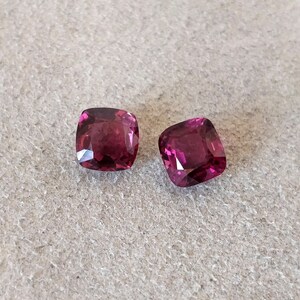 May include: Two cushion-cut gemstones, both a deep, rich purple color. The stones are set against a neutral gray background.