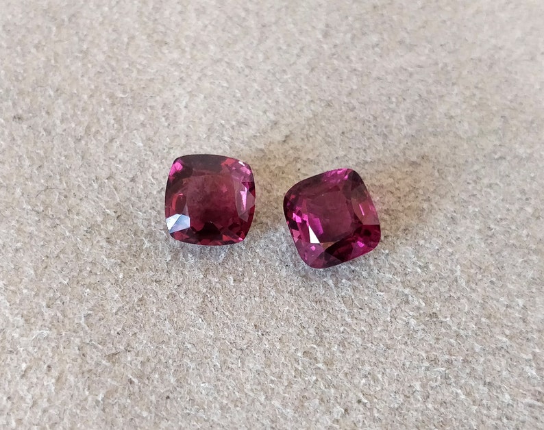 May include: Two cushion-cut gemstones, both a deep, rich purple color. The stones are set against a neutral gray background.