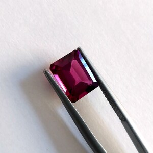 May include: A deep pink gemstone with a rectangular cut, held in tweezers against a white background.