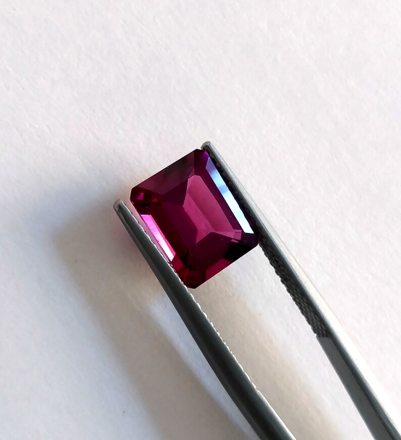 May include: A deep pink gemstone with a rectangular cut, held in tweezers against a white background.