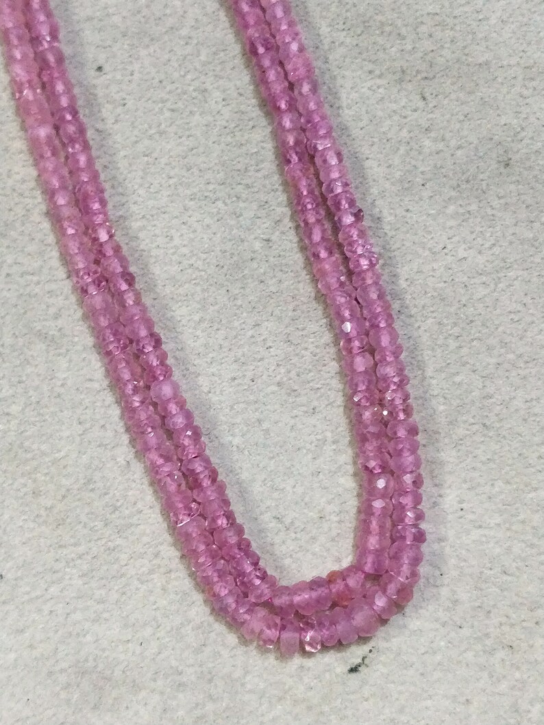 Pode incluir: Um fio de contas de pedras preciosas cor-de-rosa, provavelmente safiras cor-de-rosa, enfiadas em um fio transparente. As contas s&atilde;o facetadas e t&ecirc;m uma forma ligeiramente irregular. O fio est&aacute; dobrado ao meio, criando um formato de U.