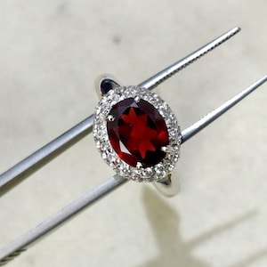 May include: A silver ring with a large, oval-shaped, red gemstone surrounded by a halo of smaller, white gemstones.