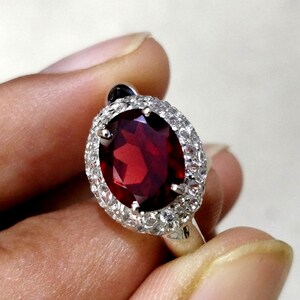 May include: A silver ring with a large oval-shaped red gemstone surrounded by a halo of smaller white gemstones.
