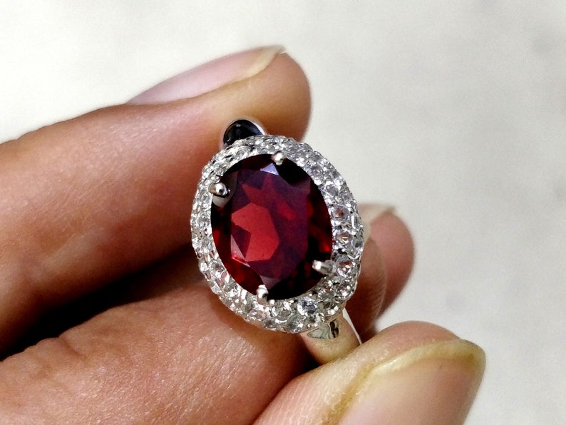 May include: A silver ring with a large oval-shaped red gemstone surrounded by a halo of smaller white gemstones.