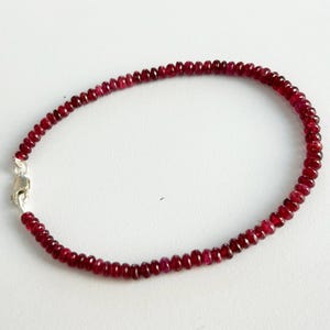 Op de afbeelding: Een delicate armband met kleine, ronde, dieprode kralen. De kralen zijn aan elkaar geregen, waardoor een doorlopende lus ontstaat, en vastgezet met een zilverkleurige sluiting. De armband wordt gepresenteerd tegen een effen witte achtergrond.