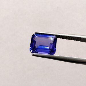 K&ouml;nnte beinhalten: Ein tiefblauer Smaragdschliff-Edelstein, der von einer Pinzette gehalten wird. Der Edelstein hat eine leuchtende blaue Farbe mit einem leichten violetten Schimmer.