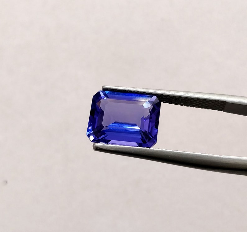 K&ouml;nnte beinhalten: Ein tiefblauer Smaragdschliff-Edelstein, der von einer Pinzette gehalten wird. Der Edelstein hat eine leuchtende blaue Farbe mit einem leichten violetten Schimmer.
