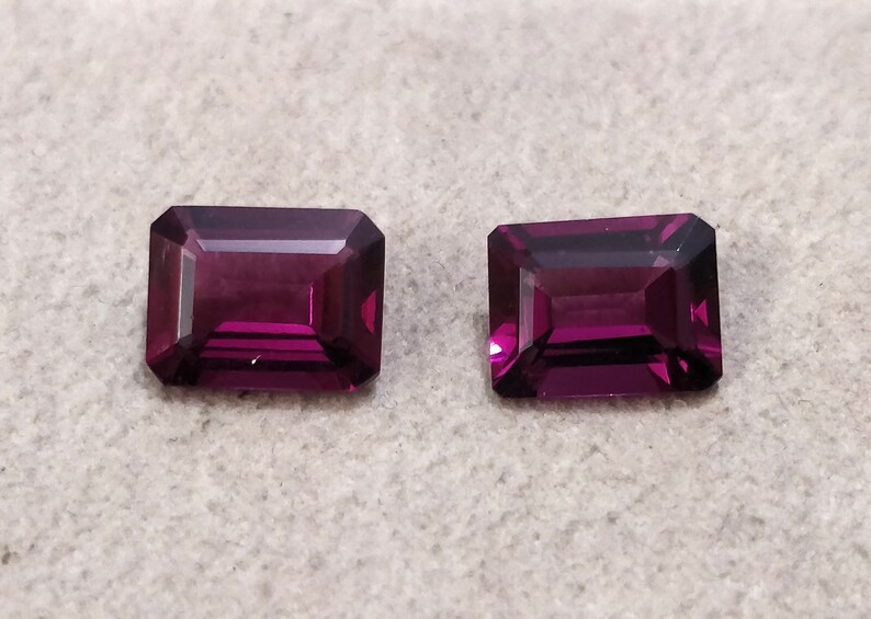 May include: Two purple rectangular gemstones with a faceted cut.