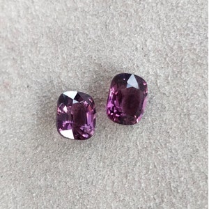 May include: Two purple cushion-cut gemstones on a gray textured surface.