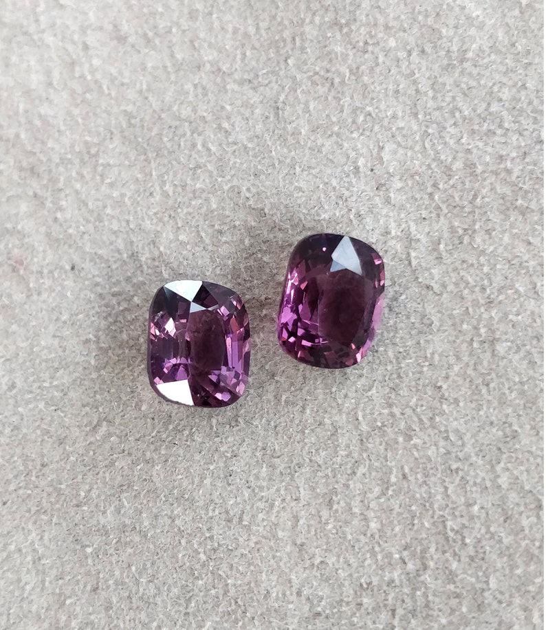 May include: Two purple cushion-cut gemstones on a gray textured surface.