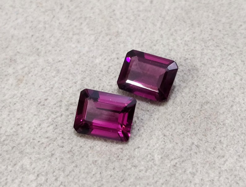 May include: Two purple rectangular gemstones with a faceted cut.