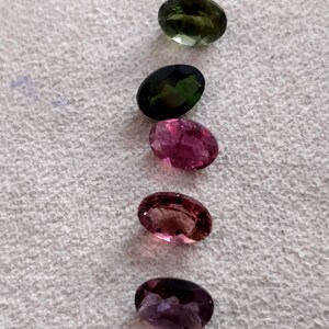 May include: Five oval-shaped gemstones in various shades of green, pink, and purple. The gemstones are arranged vertically in a row on a white surface.