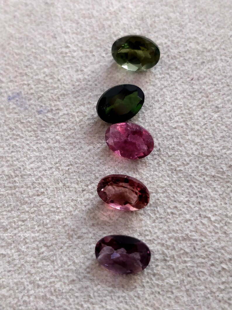 May include: Five oval-shaped gemstones in various shades of green, pink, and purple. The gemstones are arranged vertically in a row on a white surface.