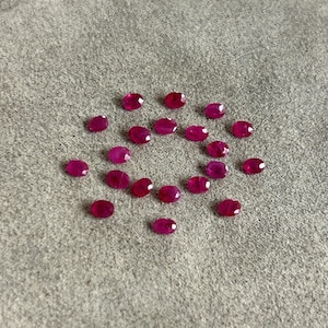 May include: A collection of nineteen small, oval-shaped, faceted rubies in a vibrant deep pinkish-red hue. Each gemstone features a polished surface with multiple facets that reflect light, arranged in a loose circular pattern on a textured surface.
