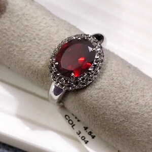May include: A silver ring with a large oval-shaped red gemstone surrounded by smaller white gemstones. The ring is on a white and gray surface with the text "COL 3.04 4.54".