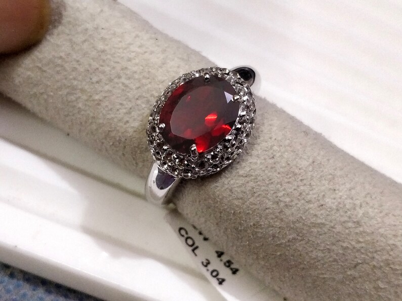 May include: A silver ring with a large oval-shaped red gemstone surrounded by smaller white gemstones. The ring is on a white and gray surface with the text "COL 3.04 4.54".