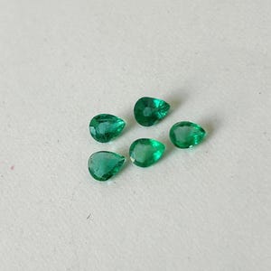 K&ouml;nnte beinhalten: F&uuml;nf lose, birnenf&ouml;rmige Smaragd-Edelsteine mit leuchtend gr&uuml;ner Farbe und facettierten Oberfl&auml;chen. Jeder Stein zeigt eine klare Tropfensilhouette mit mehreren polierten Facetten, die das Licht reflektieren und in einer kleinen Gruppe angeordnet sind.