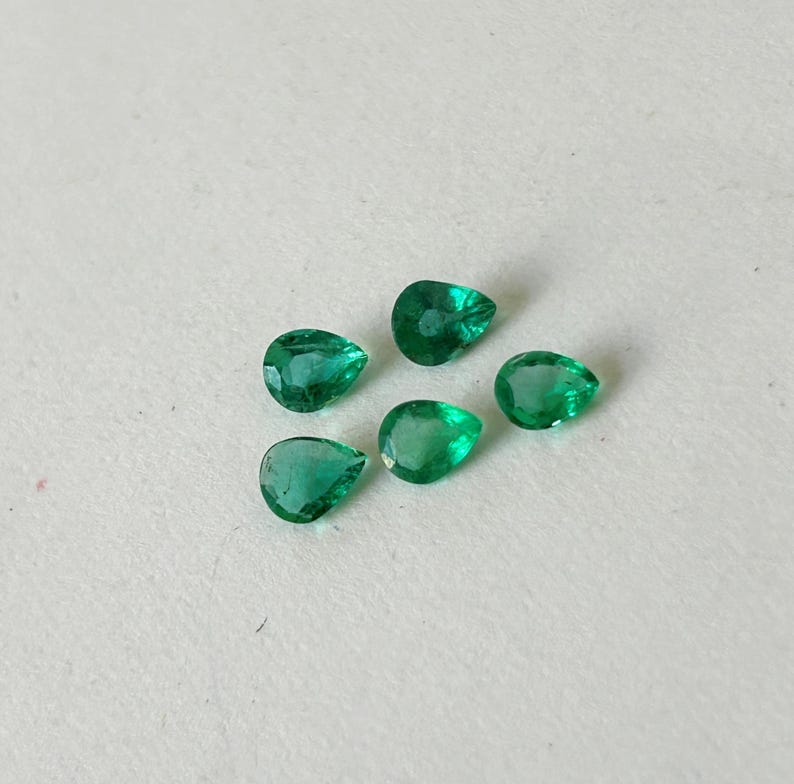 K&ouml;nnte beinhalten: F&uuml;nf lose, birnenf&ouml;rmige Smaragd-Edelsteine mit leuchtend gr&uuml;ner Farbe und facettierten Oberfl&auml;chen. Jeder Stein zeigt eine klare Tropfensilhouette mit mehreren polierten Facetten, die das Licht reflektieren und in einer kleinen Gruppe angeordnet sind.