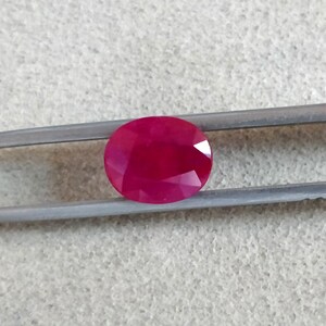 Puede incluir: Una gema ovalada de color rojo vibrante sostenida por unas pinzas. La gema es lisa y pulida, con un tono rojo intenso.