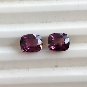 May include: Two cushion-cut purple gemstones on a white background.