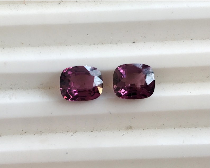 May include: Two cushion-cut purple gemstones on a white background.